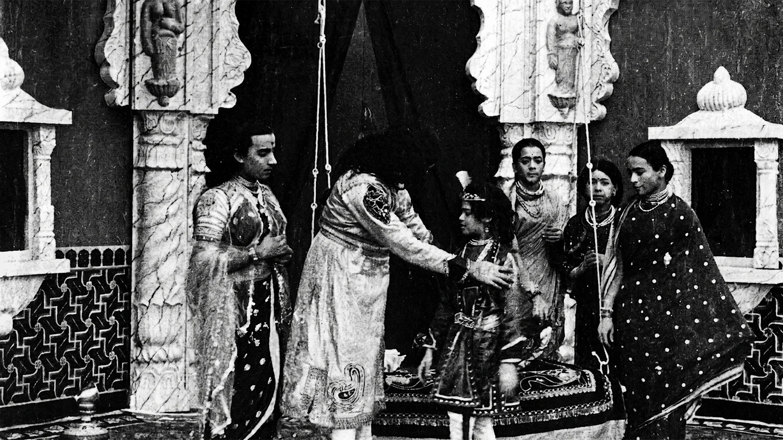 A black and white image shows several people in traditional Indian attire in an ornate indoor setting with pillars, where a man in the center places his hands on a child's shoulders.