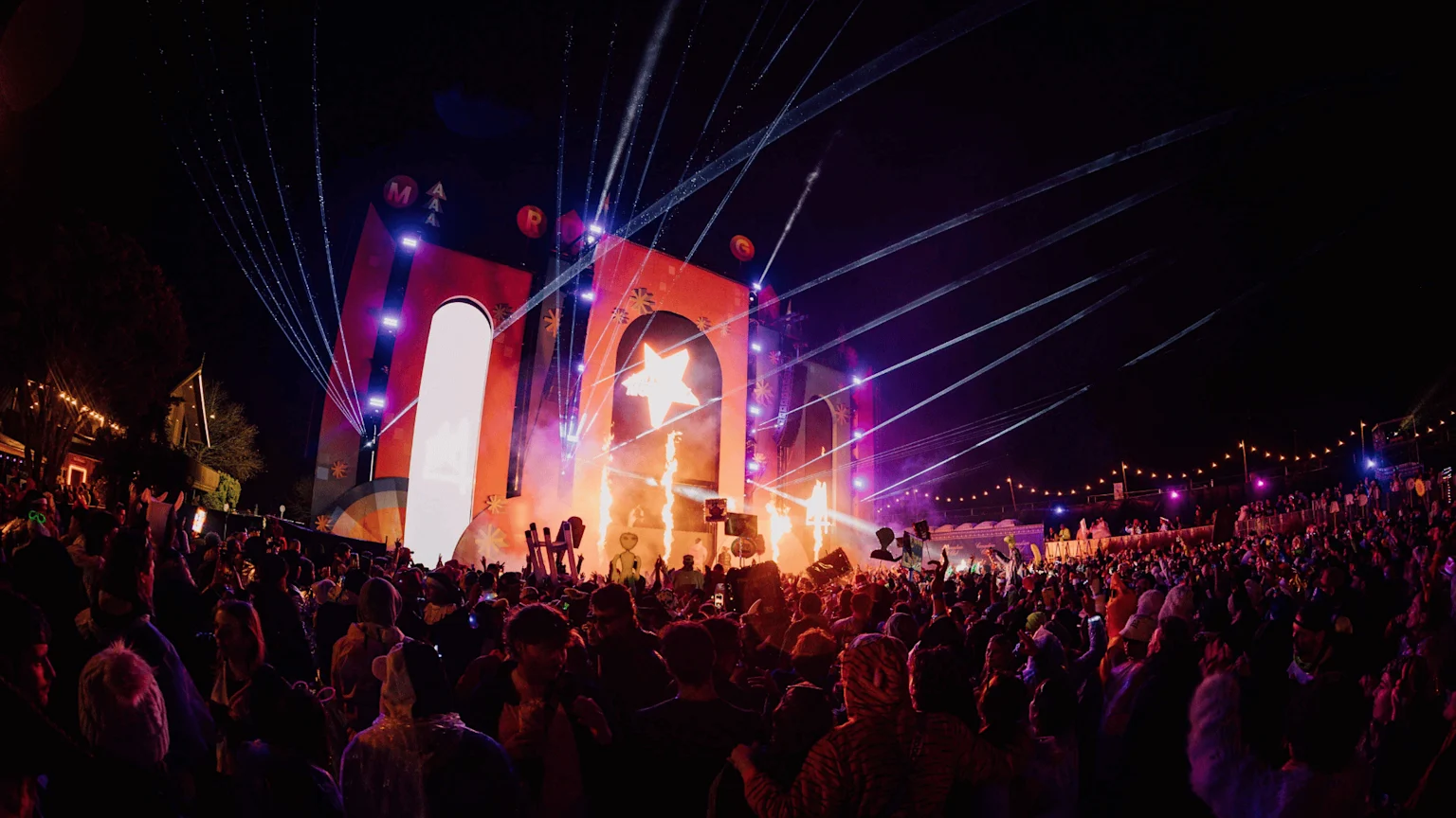 A vibrant night scene at an outdoor festival shows a large stage with a glowing star, pyrotechnics, bright lights, laser beams, and letters, surrounded by an excited crowd of people.