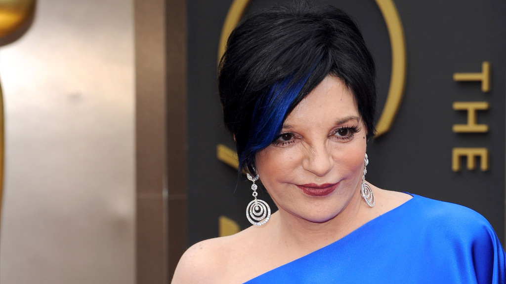 Liza Minnelli, with black hair and a blue streak, wears a blue one-shoulder dress and diamond earrings, smiling slightly against a dark background with gold elements.