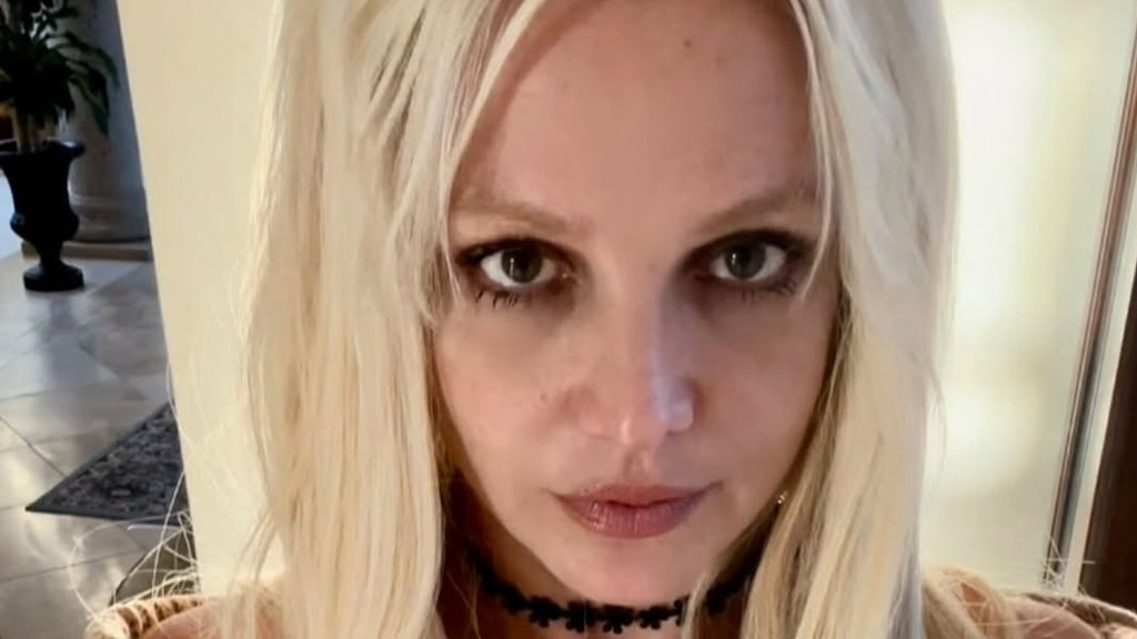 A close-up selfie of Britney Spears looking directly at the camera with dark eye makeup and a black flower-shaped choker necklace, her long blonde hair framing her face, in an indoor setting.