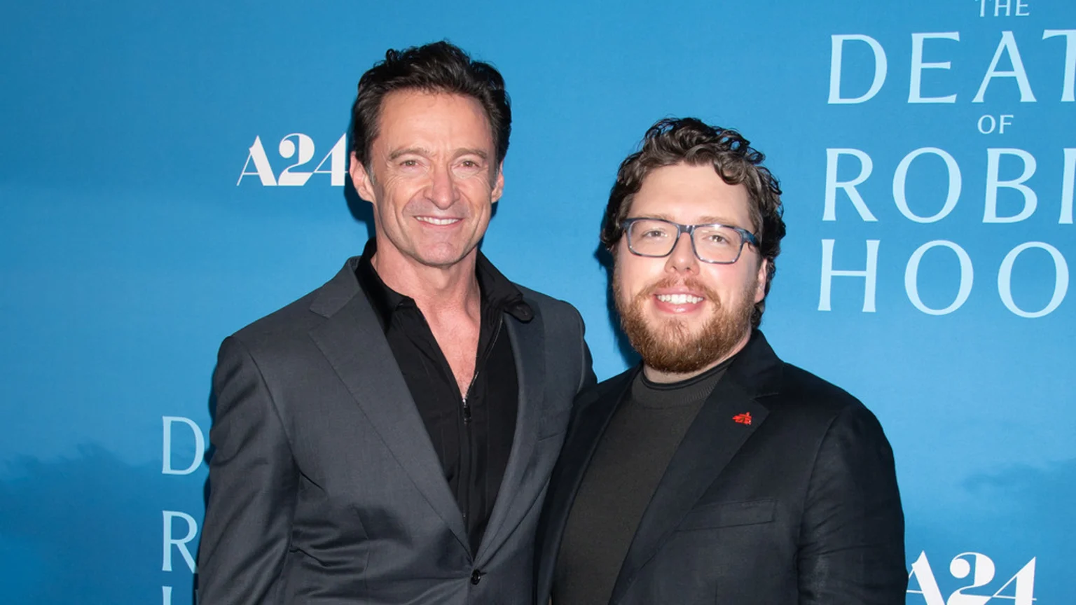 Hugh Jackman in a grey suit and Michael Sarnoski in a black blazer and glasses smile for a photo in front of a blue A24 backdrop.