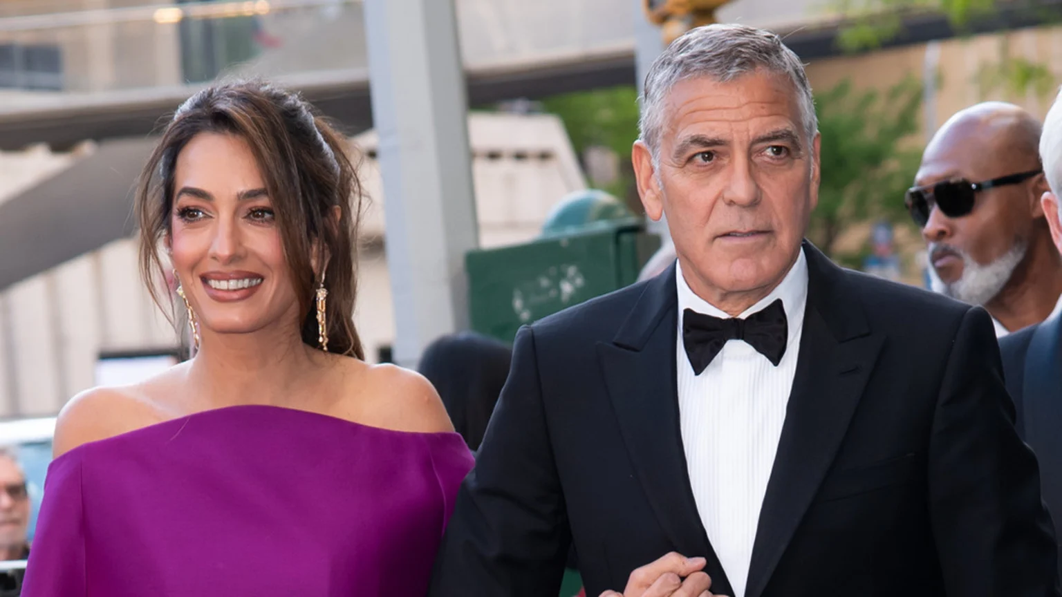Amal Clooney in a purple off-the-shoulder dress smiles while walking arm-in-arm with George Clooney, who wears a black tuxedo and bow tie.