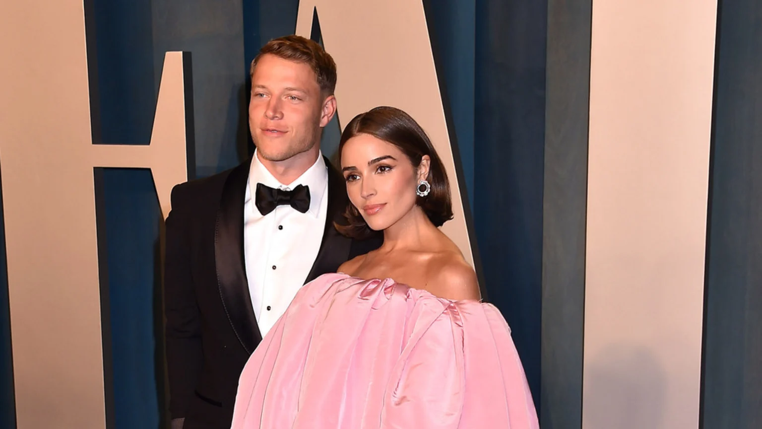Olivia Culpo in a voluminous pink off-the-shoulder gown and Christian McCaffrey in a black tuxedo pose together against a blue background.