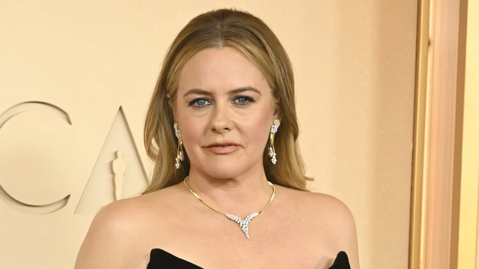 Close-up of actress Alicia Silverstone wearing a black strapless top, a gold and diamond necklace, and matching earrings, against a light tan background with "CA" and an Oscar statue cut-out.