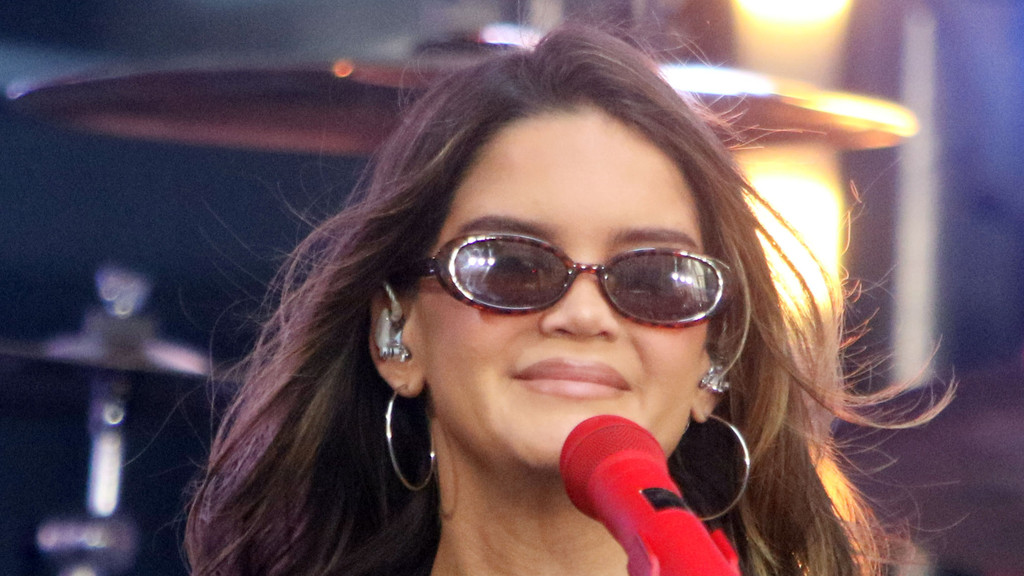 A smiling woman with long brown hair and tortoise-shell sunglasses wears in-ear monitors and hoop earrings while holding a red microphone and performing on stage.