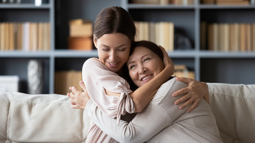 A smiling younger woman hugs an older woman warmly on a light-colored couch, expressing affection, with bookshelves visible in the background.