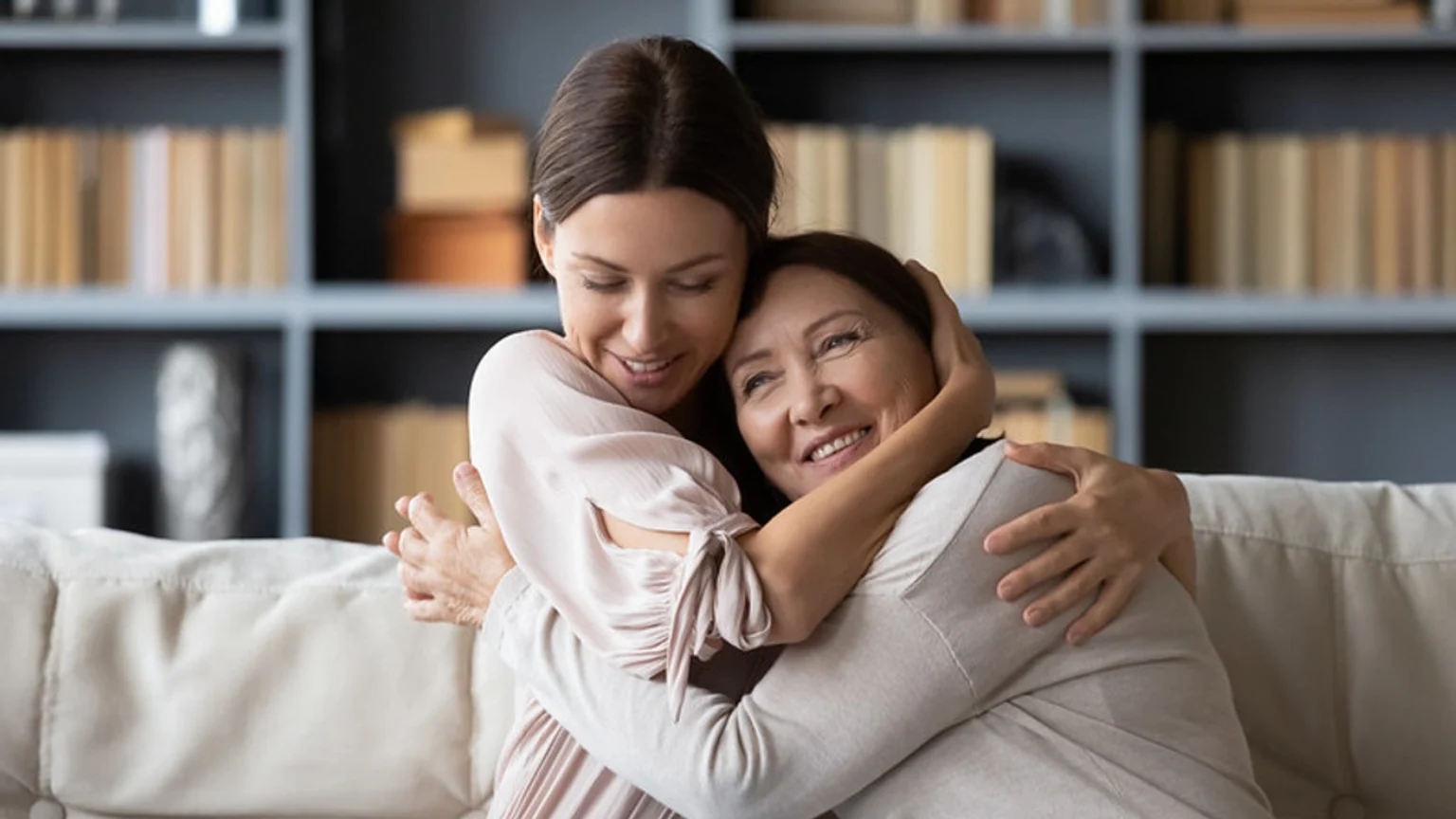 A smiling younger woman hugs an older woman warmly on a light-colored couch, expressing affection, with bookshelves visible in the background.