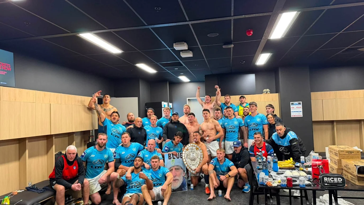 A large group of jubilant men, some shirtless and others in light blue jerseys, celebrate in a locker room, holding the Ranfurly Shield and a poster of a man's face.