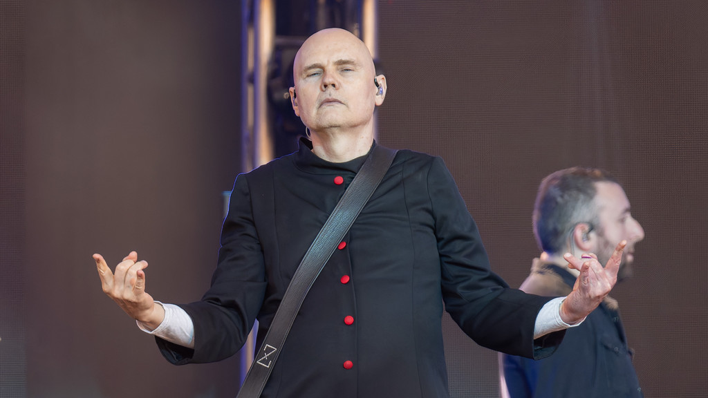 Bald musician Billy Corgan on stage, wearing a black jacket with red buttons and a guitar strap, raises his hands with a serious expression, with a blurred man in the background.