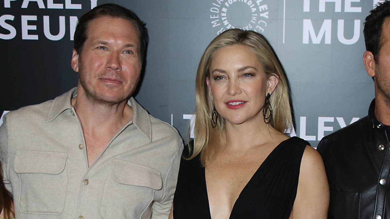 Kate Hudson, with long blonde hair in a black V-neck dress, smiles between a man in a light beige shirt and a partially visible man in a black leather jacket, against a blurred "PALEY MUSEUM" background.