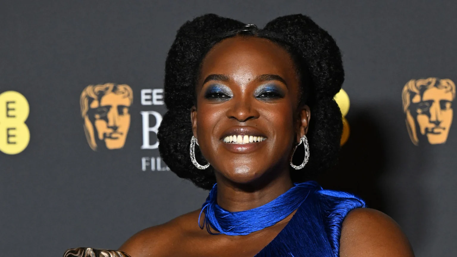 Wunmi Mosaku smiles, wearing blue eyeshadow, sparkly hoop earrings, and a blue pleated dress, with BAFTA logos visible in the dark background.