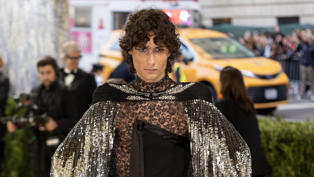 A person with curly brown hair and a serious expression wears a black lace top and a shimmering sequined cape, looking at the camera with blurred people and a yellow taxi in the background.