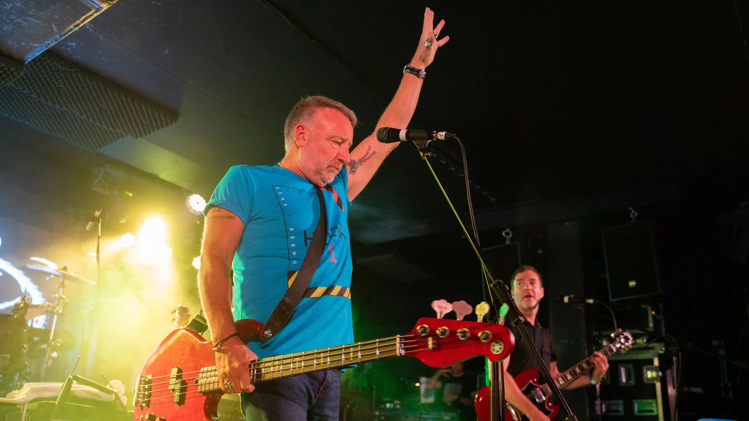 A male bassist in a blue t-shirt with a tattoo on his raised arm plays a red bass guitar on a brightly lit stage, with another guitarist visible in the dark background.