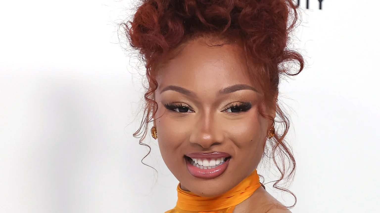 Close-up of Megan Thee Stallion smiling, with reddish-brown curly hair in an updo and tendrils, wearing a yellow top, prominent eye makeup, and glossy lips.