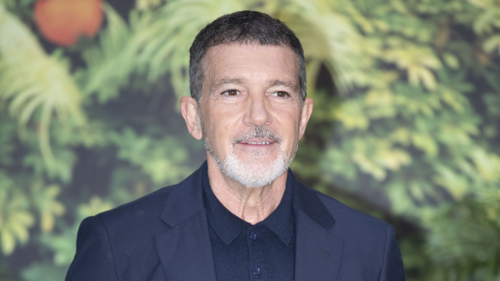 Antonio Banderas smiles slightly, dressed in a dark blue suit jacket and collared shirt, against a blurred green and orange foliage background.