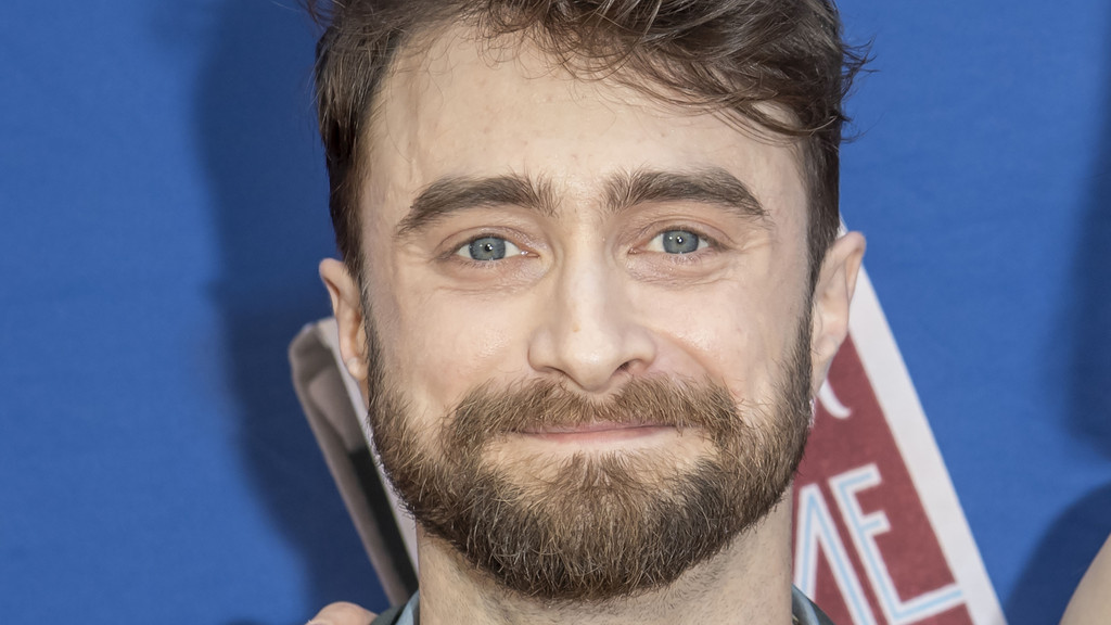 Close-up of actor Daniel Radcliffe smiling at the camera, with light blue eyes and a full beard, against a blue background with partial white and red lettering on the right.