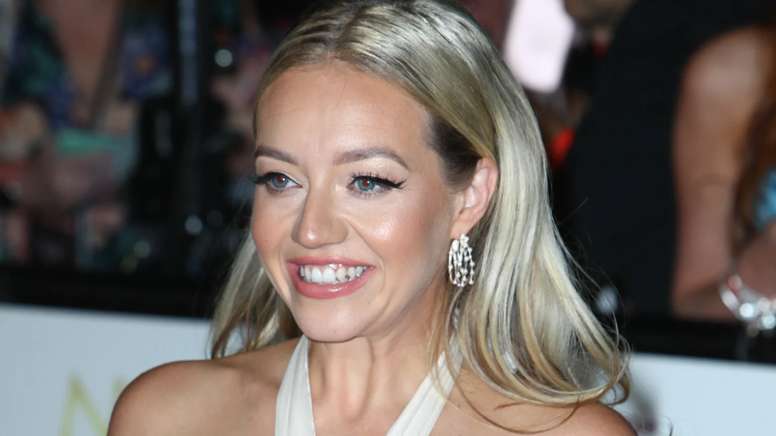 A blonde woman with blue eyes smiles brightly, showing teeth, wearing prominent eyeliner, sparkly earrings, and a light-colored halter top against a dark, blurry background.