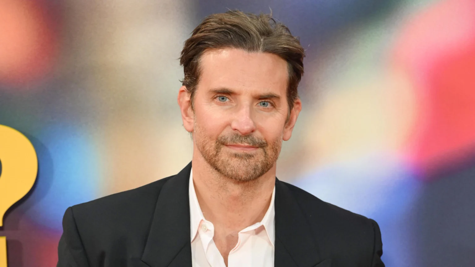 Bradley Cooper, a man with blue eyes and a beard, looks at the camera wearing a dark suit jacket and light shirt, against a blurred, colorful background with a yellow question mark.