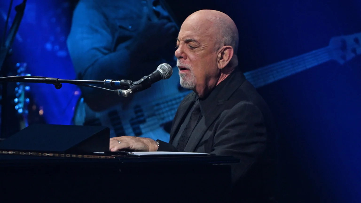Musician Billy Joel, a bald man with a goatee, sings into a microphone while playing a piano on a dark blue stage, with blurred images of instruments behind him.