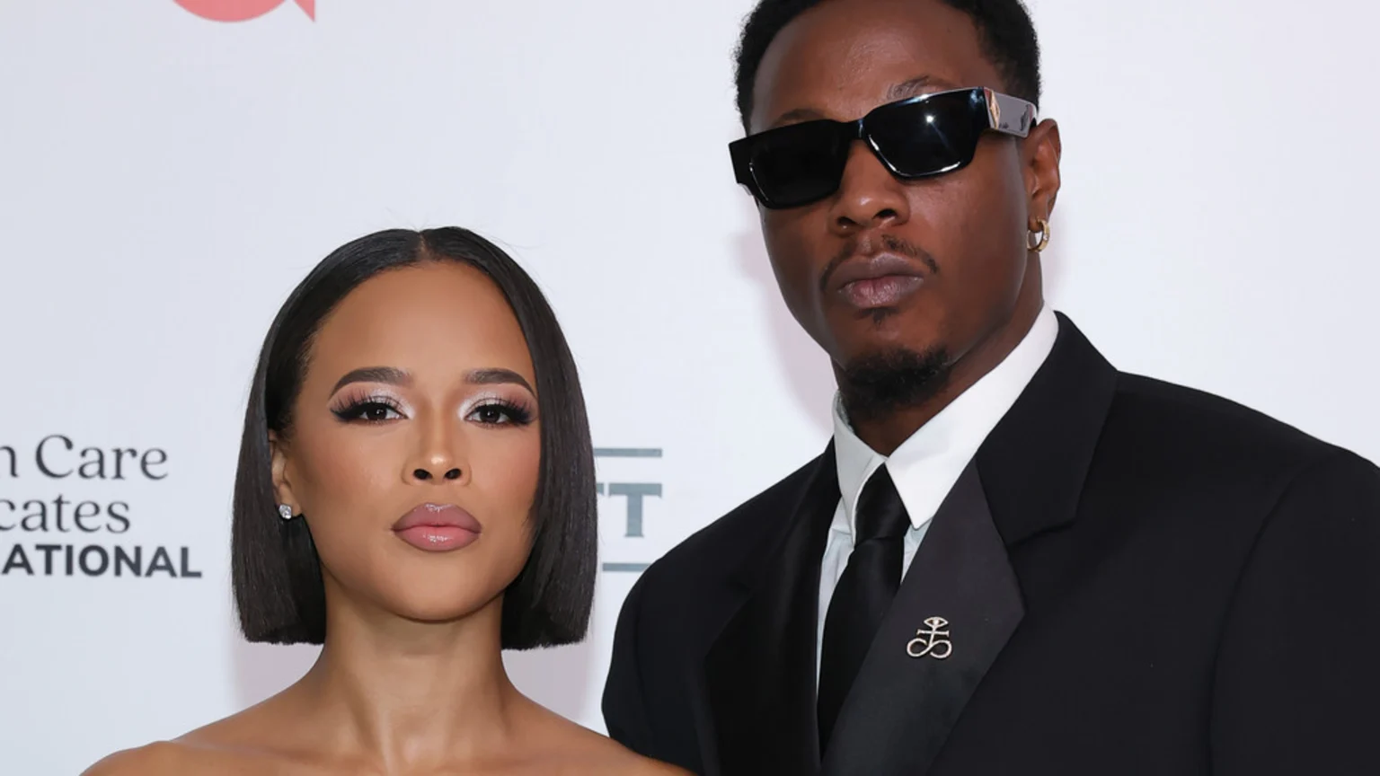A woman with a short dark bob and a man in sunglasses, a black suit, tie, and a lapel pin pose side-by-side, looking at the camera.