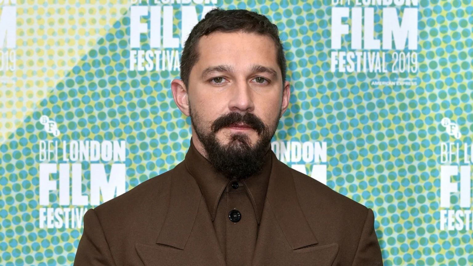 Shia LaBeouf, with a beard and mustache, wearing a brown suit, looks directly at the camera in front of a patterned backdrop for the BFI London Film Festival 2019.