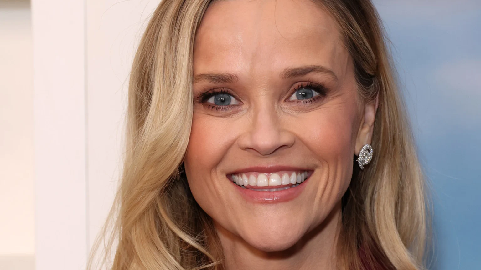 Close-up of Reese Witherspoon smiling, showing her teeth, with blonde hair, blue eyes, and a diamond earring visible on her right ear.