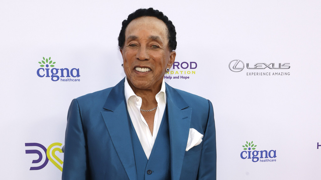 Smokey Robinson, wearing a blue suit, open-collar white shirt, and silver chain, smiles at the camera with Cigna and Lexus logos visible in the background.