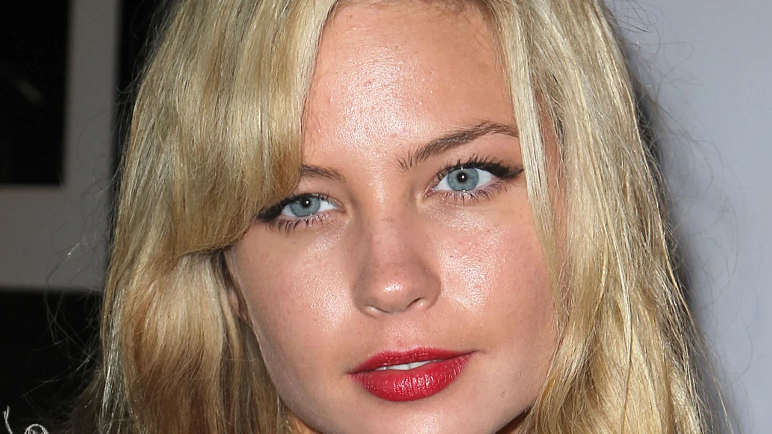 Close-up of a young woman with blonde hair, light blue eyes, and red lipstick, looking towards the camera.