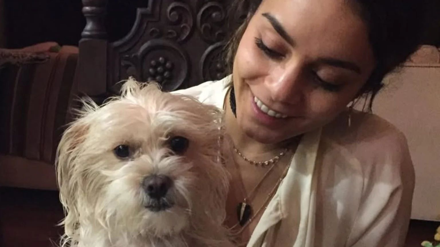 A young Vanessa Hudgens smiles warmly indoors while holding a small, fluffy white dog. She wears a light-colored collared shirt and layered gold-tone necklaces, set against a dimly lit background featuring ornate wooden furniture and brown striped fabric.