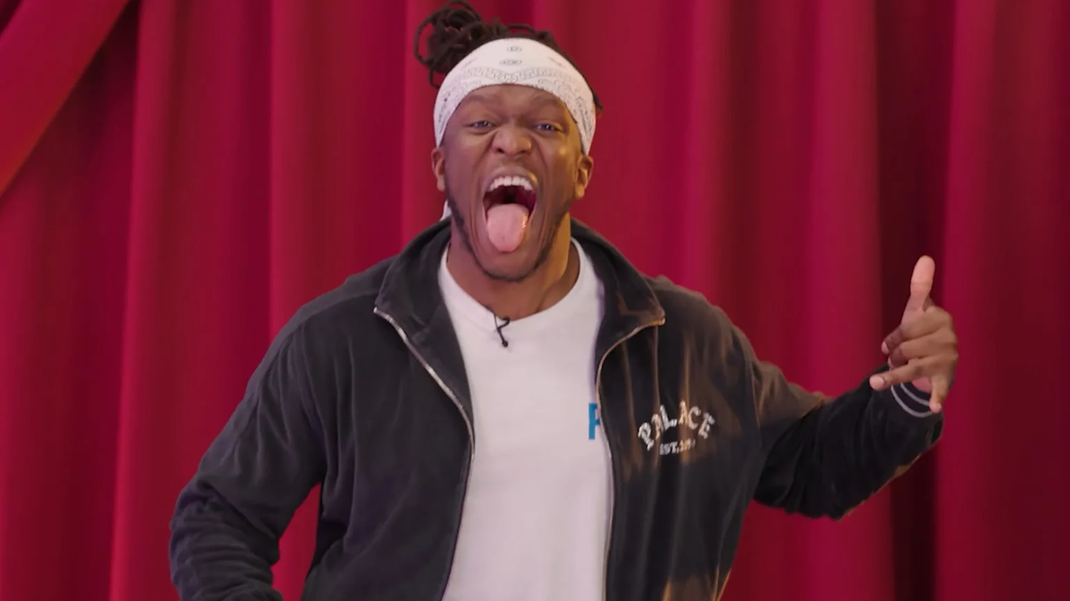 A man with a white bandana and dreadlocks intensely sticks his tongue out and widens his eyes, wearing a dark jacket over a white shirt, with a red curtain behind him. He holds his left hand up in a shaka gesture.