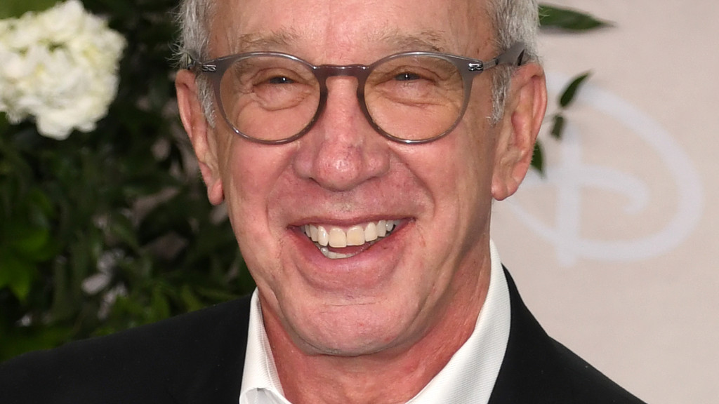 Close-up of Tim Allen smiling broadly, wearing glasses, a white shirt, and a black suit jacket, with blurred plants and a logo in the background.