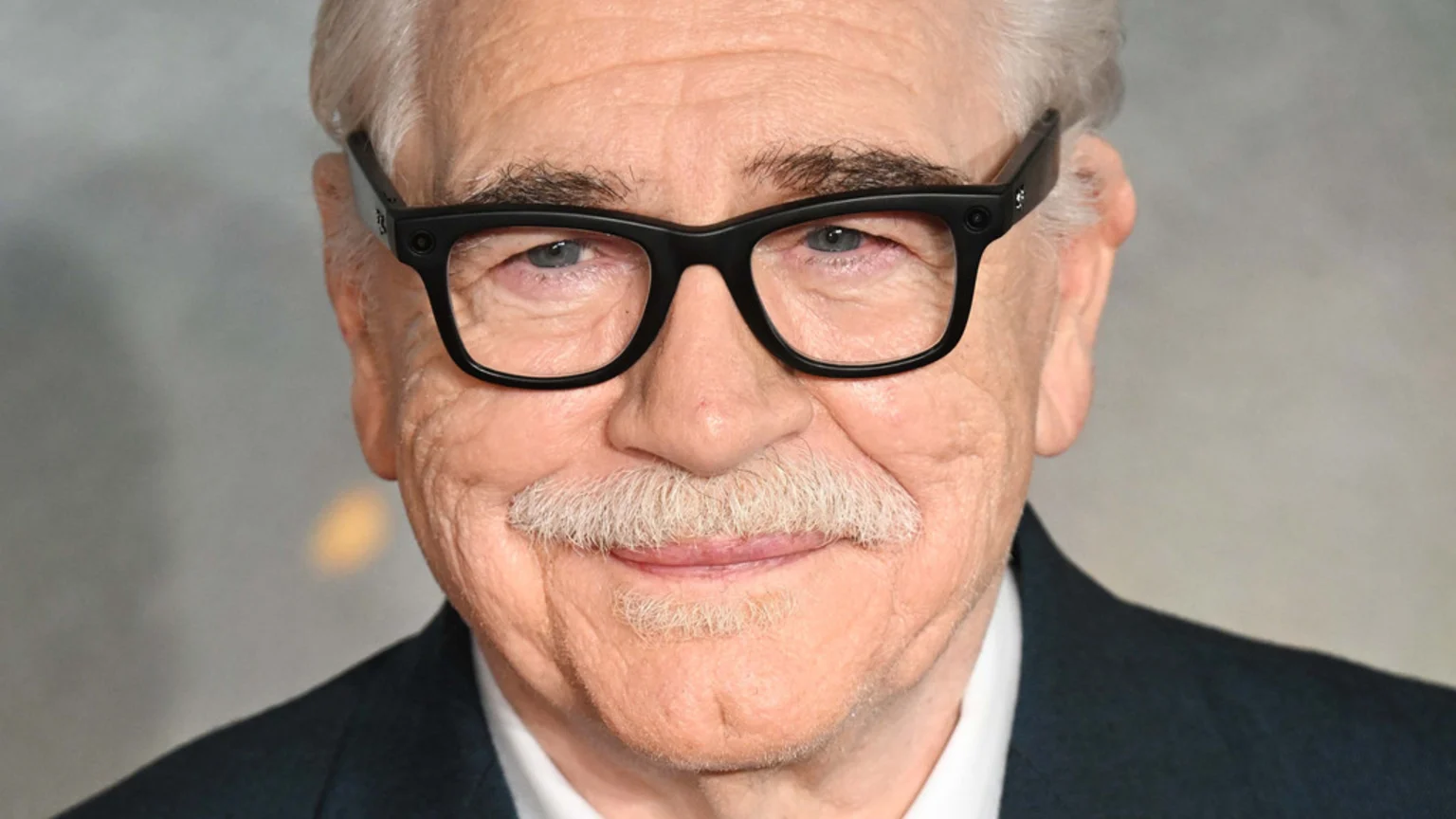 Close-up headshot of an older man with white hair, a white mustache, black square glasses, and a dark suit jacket, looking at the camera with a slight smile.