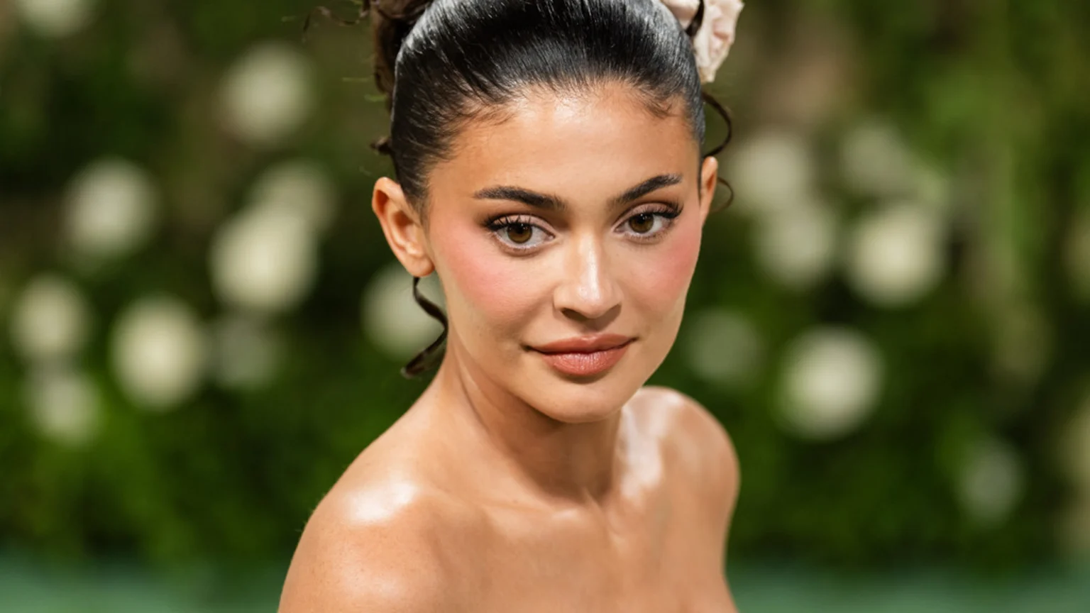 Kylie Jenner looks directly at the camera in a close-up, with her hair styled up, wearing noticeable pink blush and a nude lip against a blurred green background.
