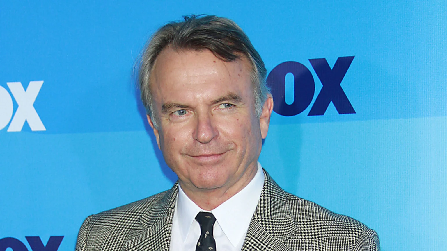 A smiling man with grey-streaked hair and blue eyes wears a houndstooth jacket and tie against a blue background with FOX logos.
