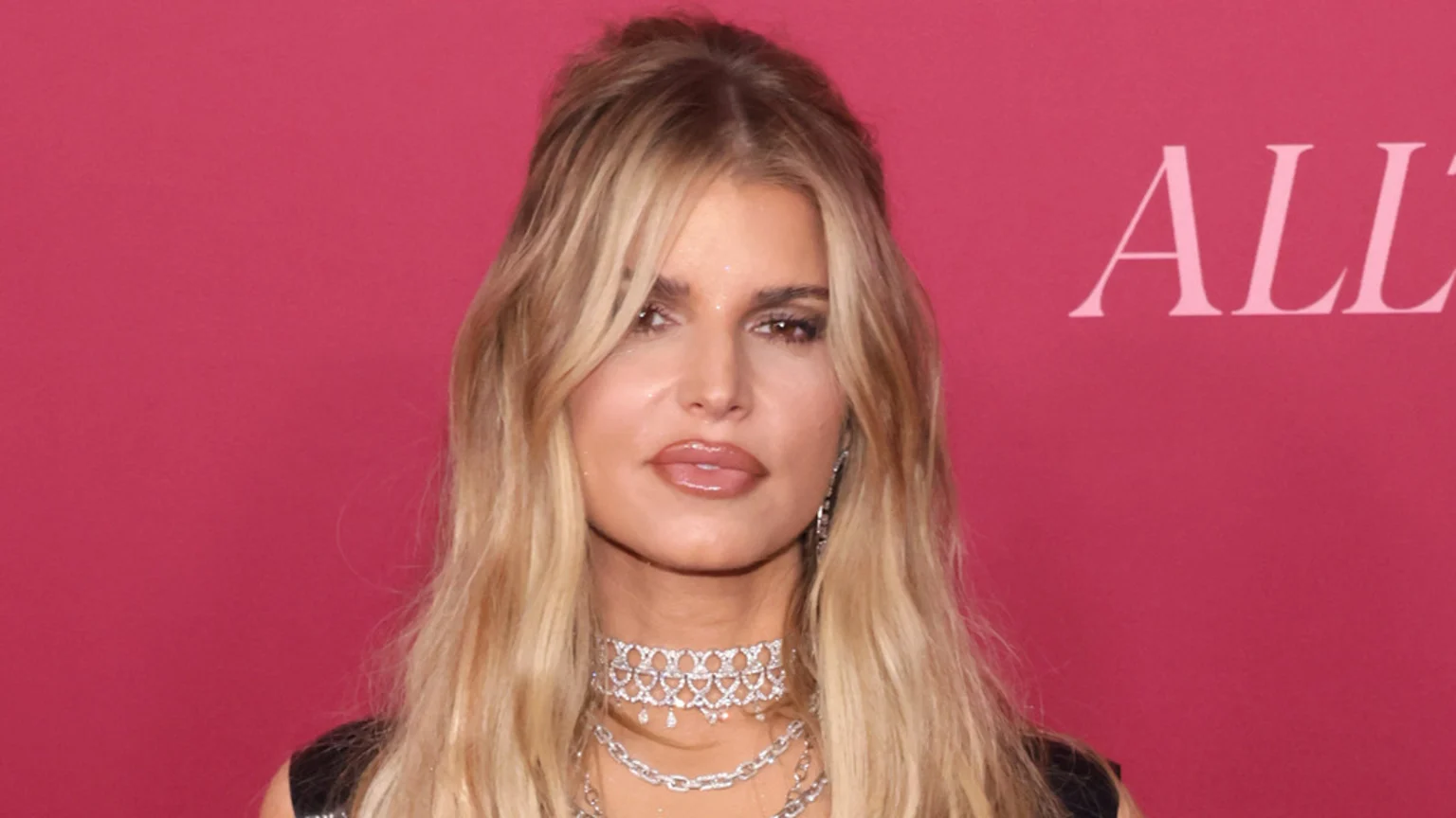 Close-up of Jessica Simpson with long blonde hair, a serious expression, and multiple sparkly necklaces against a magenta background with partial text "ALL" visible.