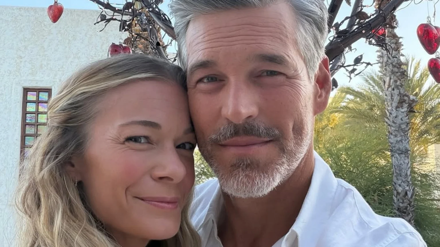 LeAnn Rimes and Eddie Cibrian smile closely together outdoors, with Rimes having blonde hair and Cibrian gray hair and a beard, against a backdrop of a light building, palm trees, and hanging red heart decorations.