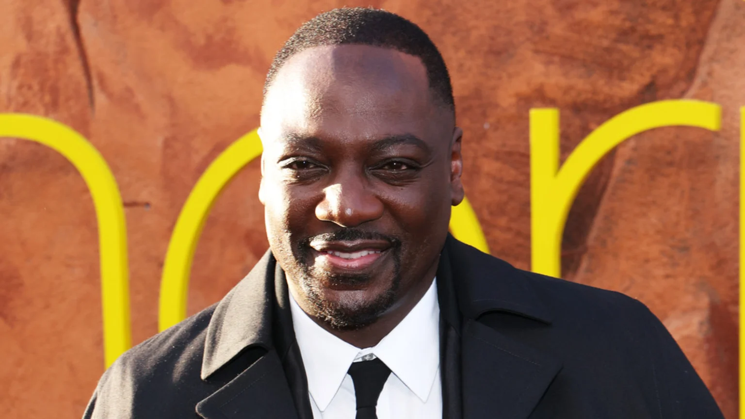 Close-up of a smiling Black man with short hair and a beard, wearing a dark coat, white shirt, and black tie against a blurred orange and yellow background.