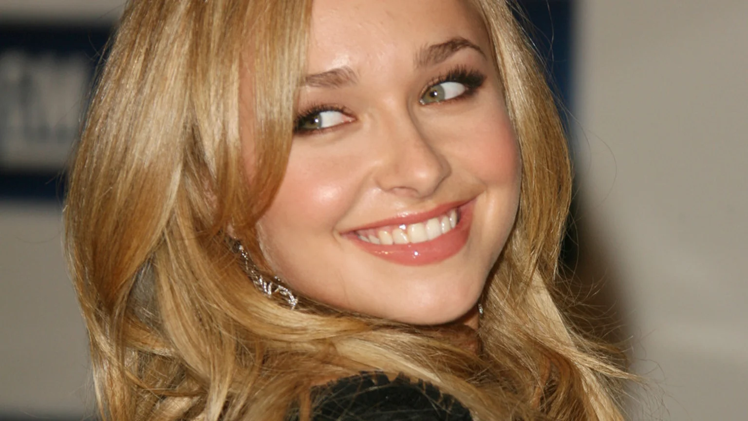 A smiling young woman with long blonde wavy hair and green eyes looks sideways, wearing glossy pink lipstick and an earring.