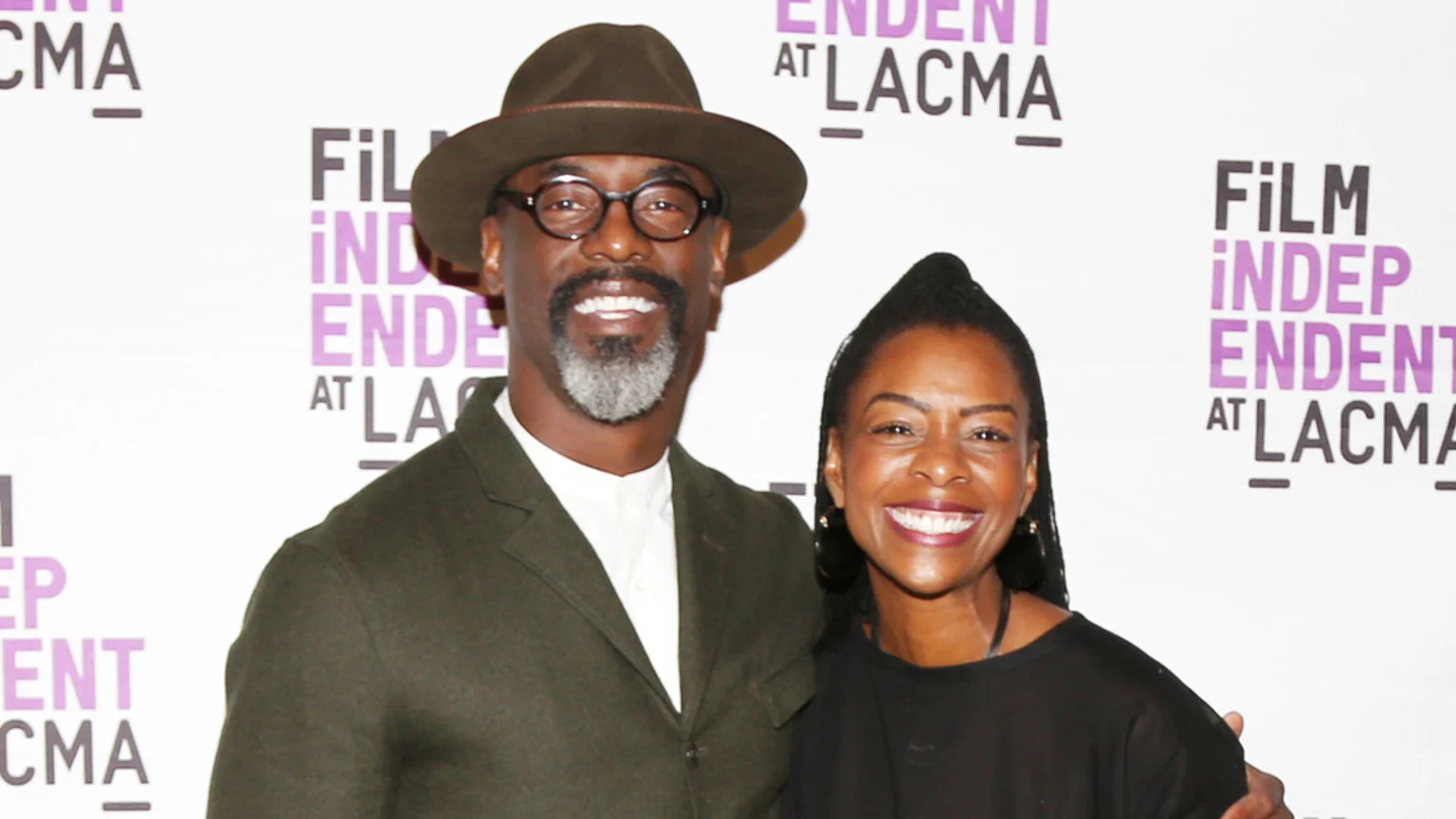 A smiling man in a hat and glasses with a gray beard stands next to a smiling woman with dark braided hair, both in front of a white banner with "FILM INDEPENDENT AT LACMA" text.
