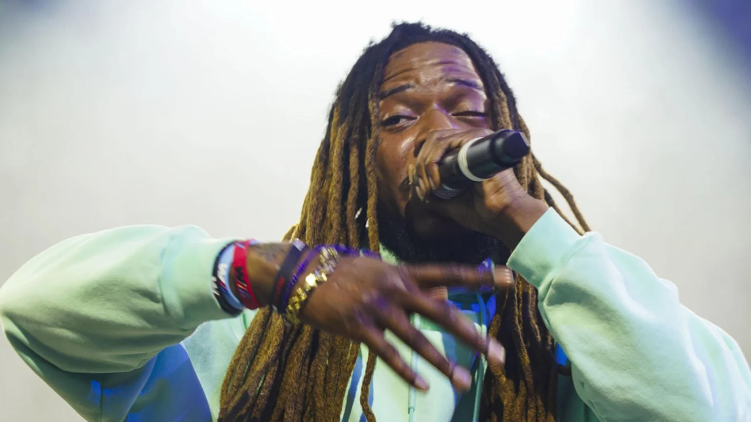 A person with dreadlocks and bracelets sings into a microphone, wearing a light green sweatshirt, against a bright, hazy background.