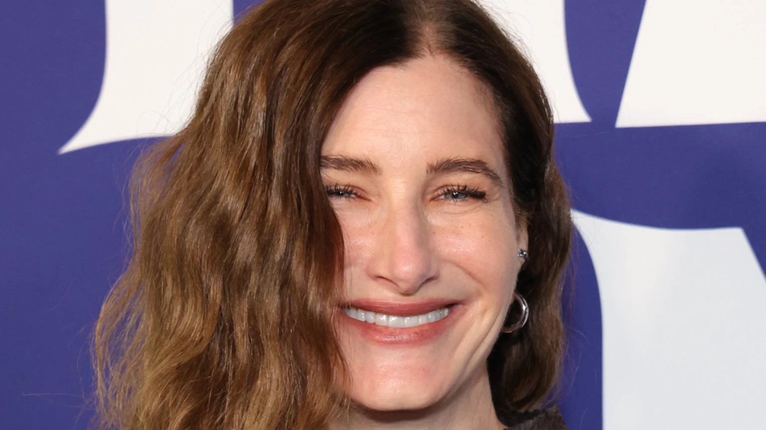 A close-up of a smiling woman with long brown wavy hair partially covering her face, blue eyes, and visible laugh lines, against a blurred purple and white background.