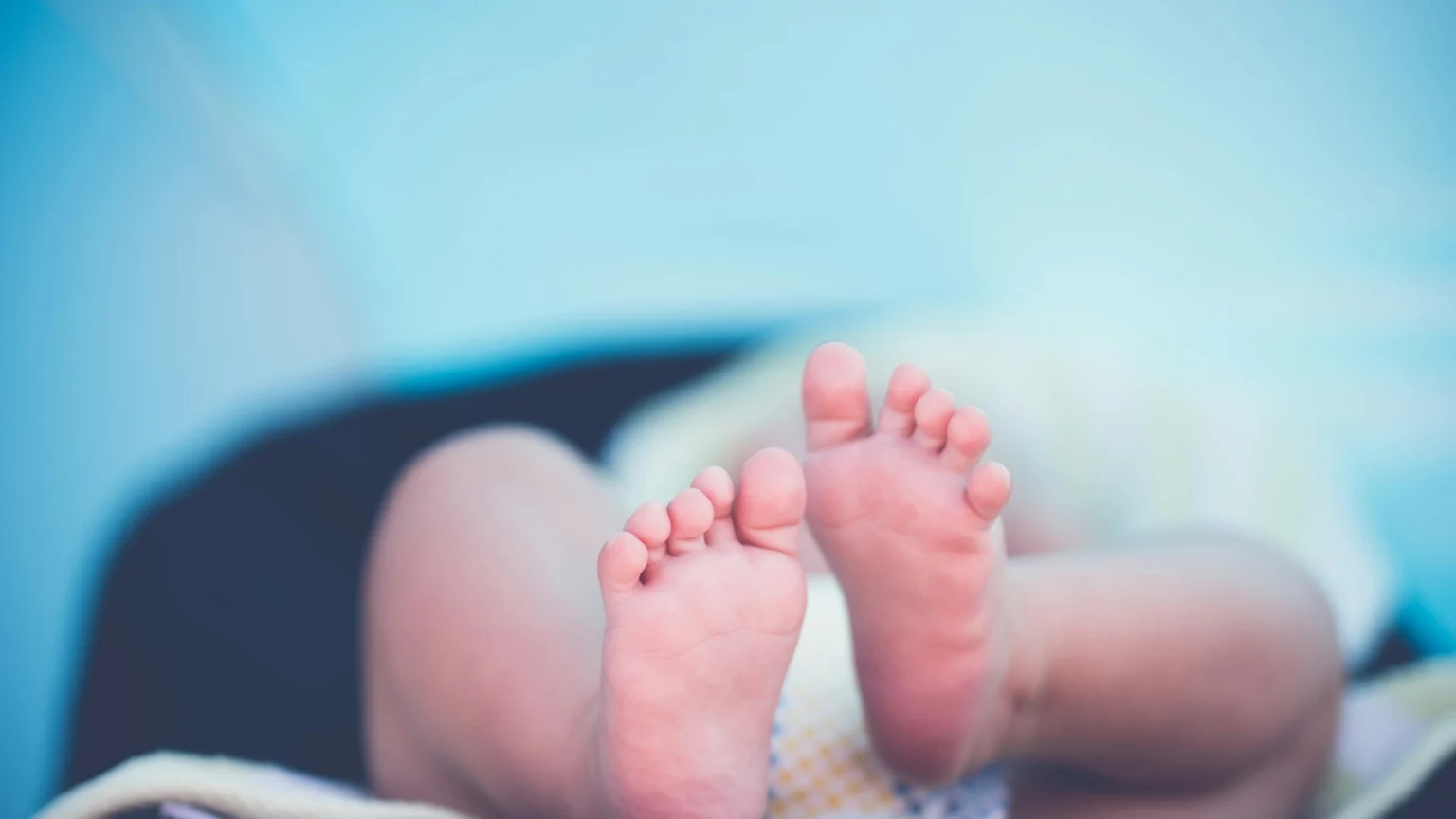 A close-up view of a baby's bare feet and curled toes, with a glimpse of a patterned diaper, against a blurred light blue background.