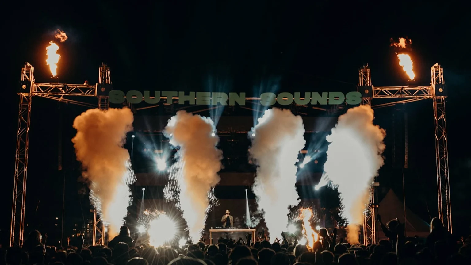 A vibrant night-time music festival scene with a DJ on stage under a "SOUTHERN SOUNDS" sign, surrounded by bright lights, pyrotechnic flames, smoke, and sparks, with a large crowd raising hands.