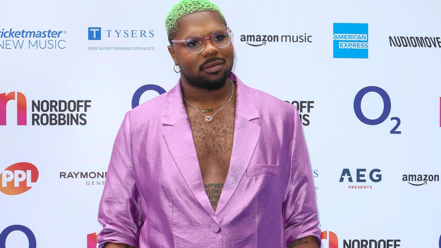 MNEK with bright green hair, glasses, and an open shiny purple blazer poses against a white backdrop featuring various sponsor logos.