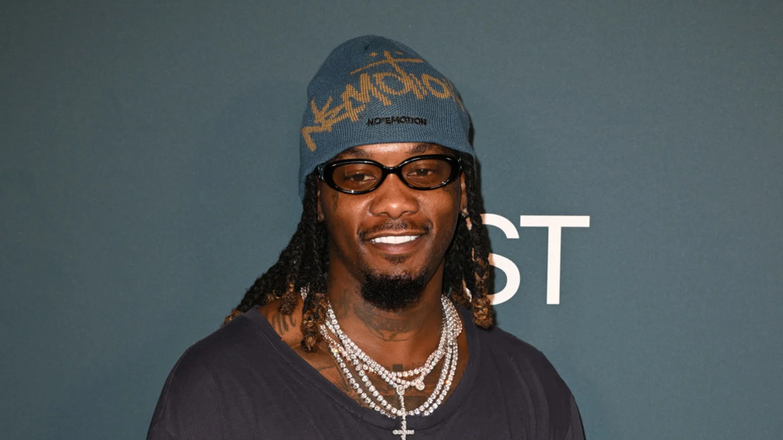 Headshot of rapper Offset smiling, wearing a blue beanie, dark glasses, and multiple diamond chains against a dark teal background.