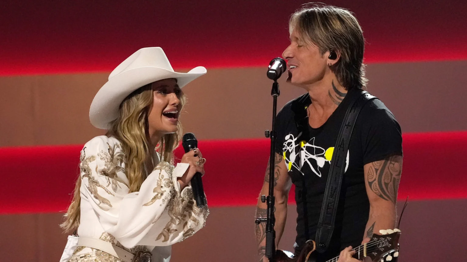 Lainey Wilson in a white cowboy hat and embroidered top sings into a microphone next to Keith Urban playing guitar and singing, against a red and gray striped background.