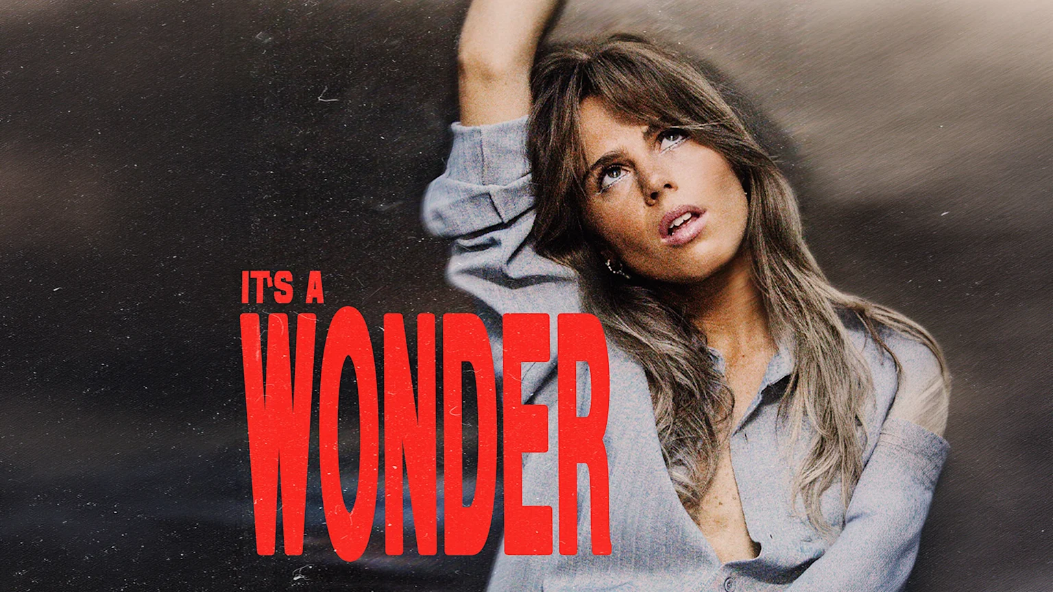 A woman with long hair in a light blue shirt looks up with her arm raised, beside large red text that reads "IT'S A WONDER" on a dark, textured background.