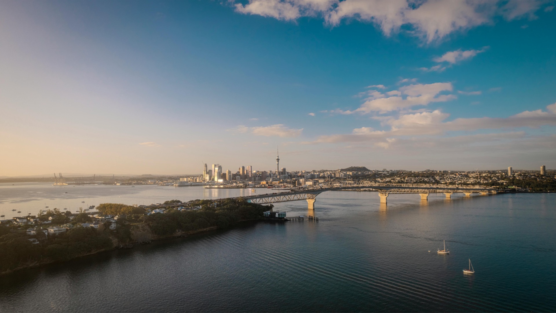 An aerial view of Auckland at sunset, featuring the city skyline with the Sky Tower, the Auckland Harbour Bridge, and boats on the water, with clouds in the sky.