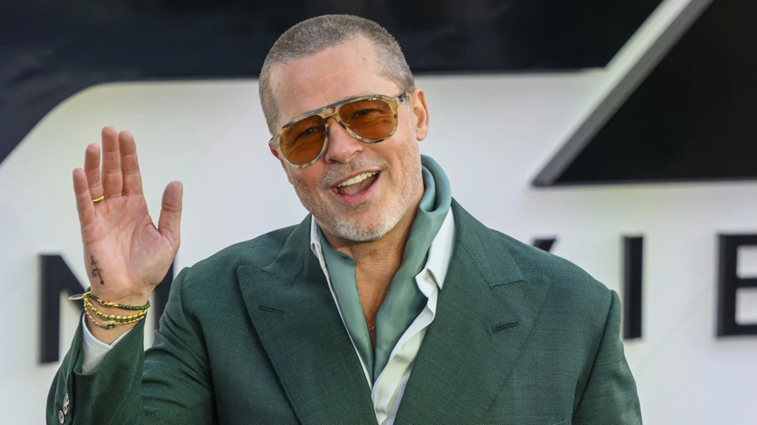 Brad Pitt smiles and waves at the camera, wearing a green suit, a light blue scarf, and amber-tinted sunglasses, with a short haircut and beard.