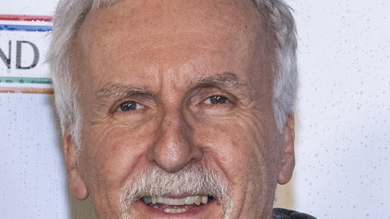 Close-up of James Cameron, a smiling older man with grey hair, mustache, and beard, looking at the camera against a light, speckled background.